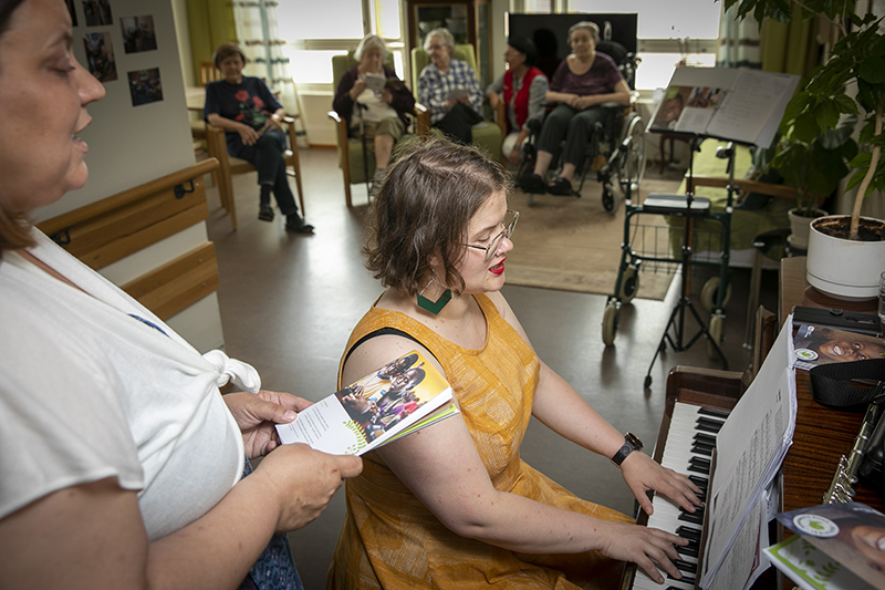 Petra Perttula laulaa ja Eveliina Ylikoski sekä laulaa että soittaa pianoa. Hoivakodin asukkaita taustalla kuuntelemassa.