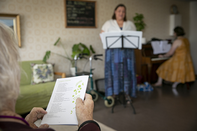Fiiliskuva. Taustalla Petra Perttula laulaa ja Eveliina Ylikoski soittaa pianoa. Kuvan etualalla hoivakodin asukkaan kädet pitelevät lauluvihkoa.