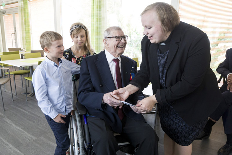 Vanha mies, joka istuu pyörätuolissa, sekä pastori kättelevät. Molemmat pitelevät samaa kirjekuorta. Takana on kaksi lasta. Mies ja pastori nauravat ja lapset hymyilevät. Tila on avara ja valoisa, siinä on pöytiä ja vihreitä tuoleja sekä isot ikkunat. Kuvan reunassa näkyy osa toisen vanhan miehen vartalosta.