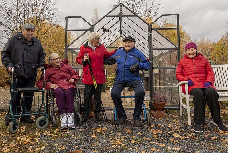 Viisi iloista vanhusta kasvihuoneen edessä ulkona.