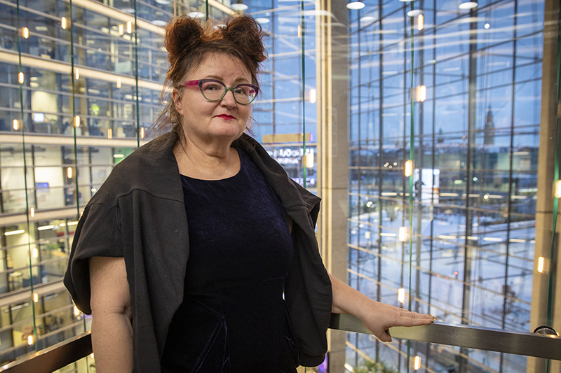 Silmälasipäinen nainen hymyilee ja seisoo ison, avaran rakennuksen ylemmässä kerroksessa ja pitää toista kättä kaiteen päällä. Takana näkyy valtavamäärä rakennuksen ikkunoita.