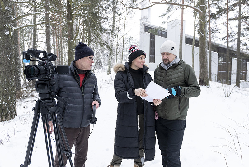 Mies ja nainen lukevat papereita. Kuvaaja katsoo kameran vierestä miestä ja naista. Talvinen maisema. Taustalla rakennus ja puita.