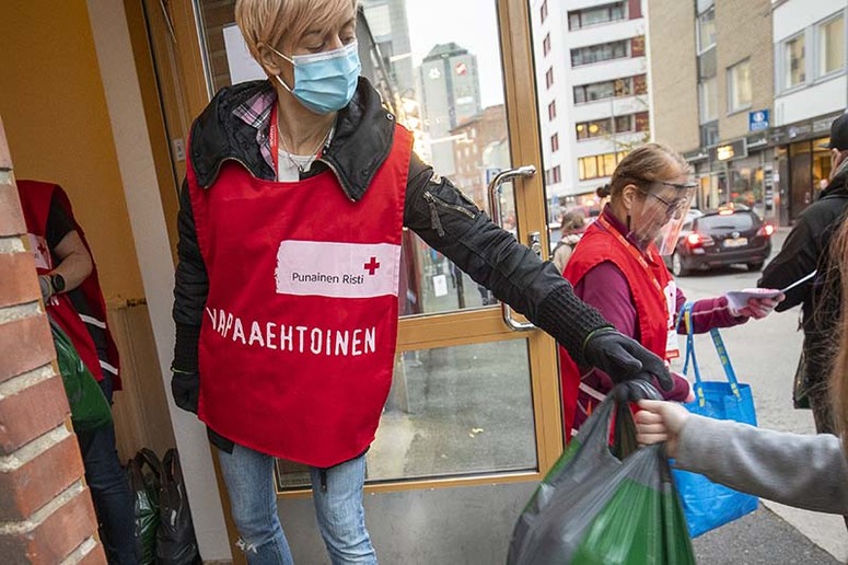 Punaiseen liivin pukeutunut vapaaehtoistyöntekijä antaa ruokakassia oven suusta ihmiselle.