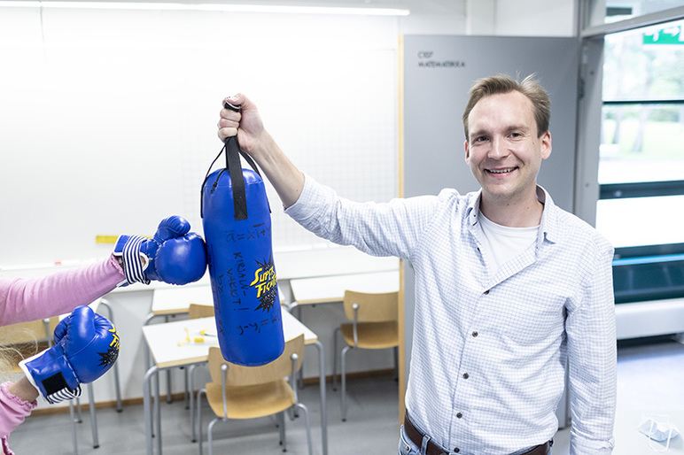 Hymyilevä henkilö pitelee minikokoista nyrkkeilysäkkiä, ja kuvan reunasta tulee kaksi kättä nyrkkeilyhanskoissa.