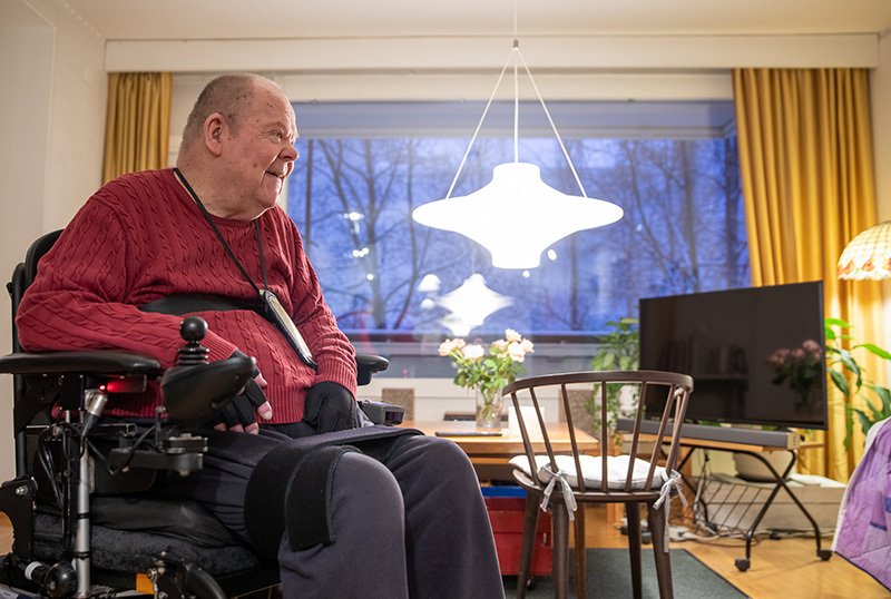 Mies sähköpyörätuolissa etualalla, taustalla näkyy huone ja huonekaluja.