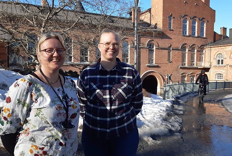 Kaksi naista keväällä kaupungissa sisävaatteissa teollisuusympäristössä. Taustalla joku pyöräilee.