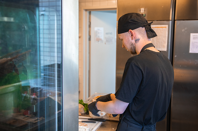 Mustapaitainen mies puolittaa leipää käsillään keittiössä. Hänellä on yllään musta kokinhattu sekä mustat kumihanskat.