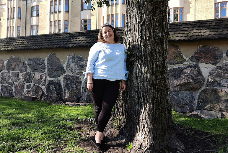 Henkilö nojaa puun runkoon. Hänen takanaan aurinkoisessa maisemassa on korkea aita ja sen takana kerrostaloja.