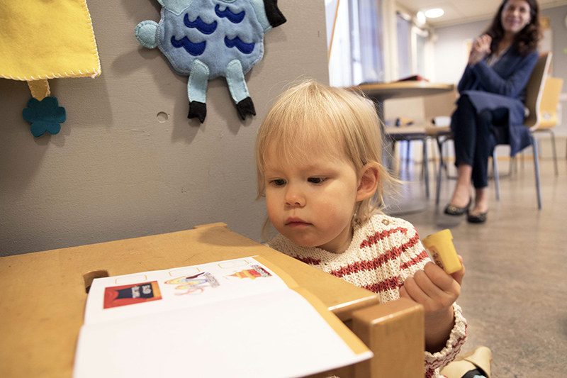 Pieni lapsi istuu vihkon edessä, esine kädessään. Taustalla istuu aikuinen.