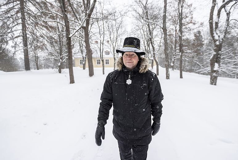Tummiin vaatteisiin pukeutunut mies kulkee lumivalkoisessa maisemassa. Taustalla on puita.