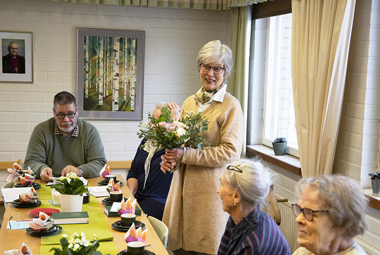 Kahvipöydän ääressä on ihmisiä, joista yksi seisoo kukkakimppu kädessään.
