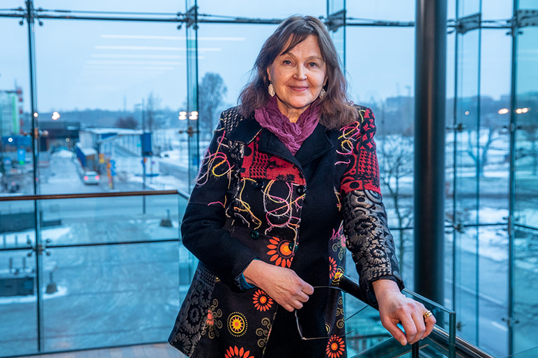 Nainen nojaa kaiteeseen ja hymyilee kameralle. Hänen takanaan näkyy valtavat ikkunat ja talvinen kaupunkimaisema.