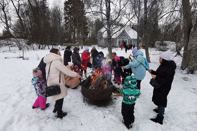 Ulkona lumisessa maisemassa on pata ja siinä nuotio. Lapset ja aikuiset pitävät käsissään keppejä, jotka osoittavat nuotioon.