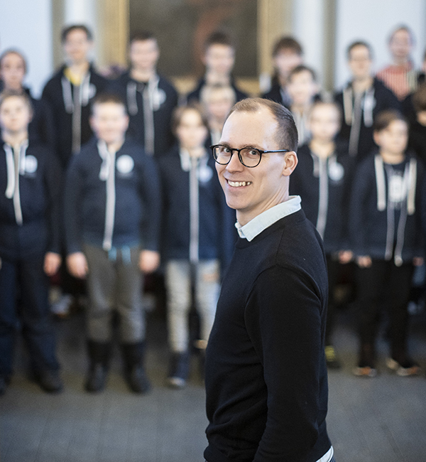 Silmälasipäinen mies on puoliksi kääntyneenä katsojaan päin. Taustalla on ryhmä nuoria poikia, joilla on samanlaiset takit.