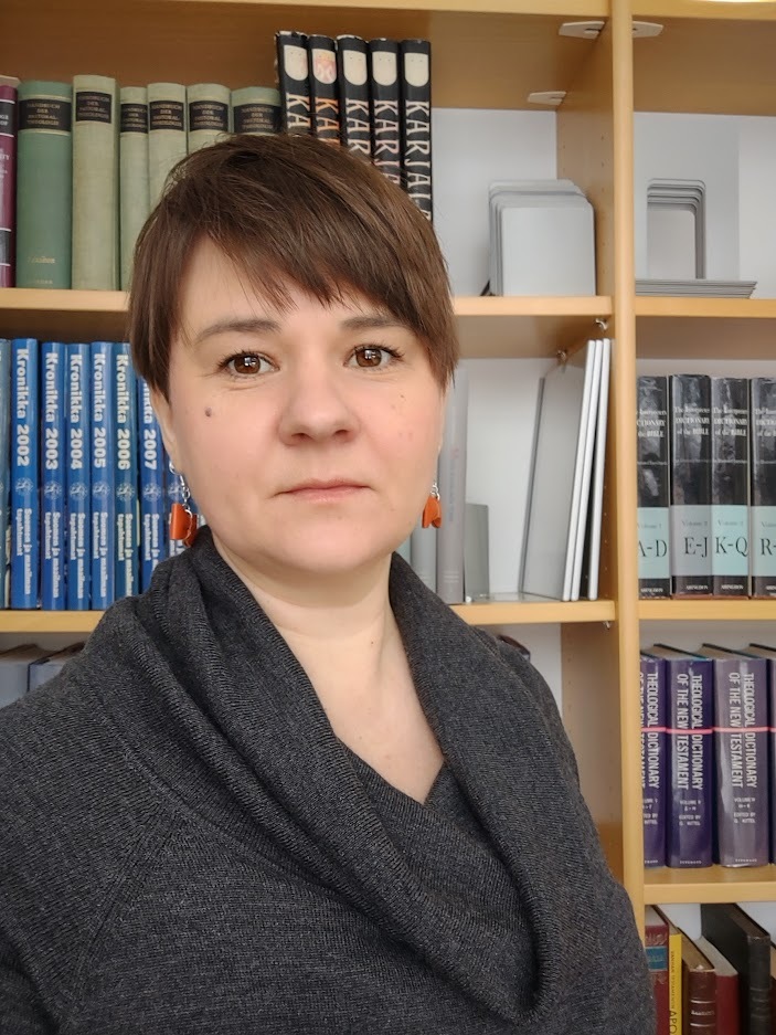 Henkilökuvassa keski-ikäinen lyhythiuksinen nainen seisoo kirjahyllyn edessä pieni hymy kasvoillaan.