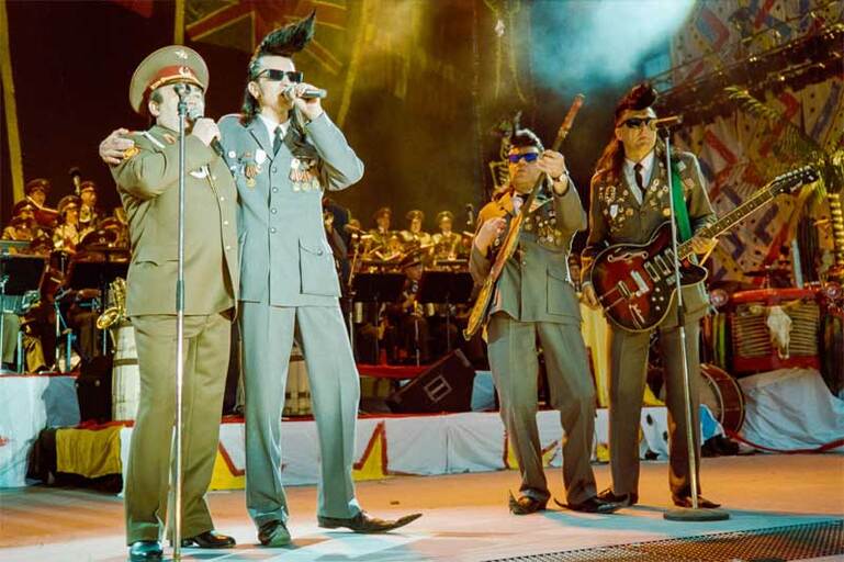Leningrad Cowboys hiustyylin omaava henkilö pitää kättä Puna-armeijan uniformuun puketuneen miehen olkapäällä, taustalla kaksi kitaristia, joilla on Puna-armeijan uniformu.