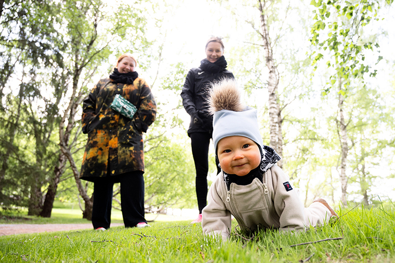 Vauva ryömii vihreällä nurmella kohti. Aikuiset katselevat taustalla.