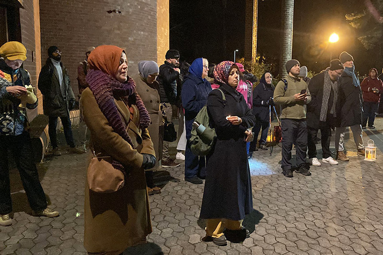 Ihmisiä seisoo syksyisissä vaatteissa ulkona pimeän aikaan laatoituksen päällä rakennuksen vieressä. Lyhdyt valaisevat.