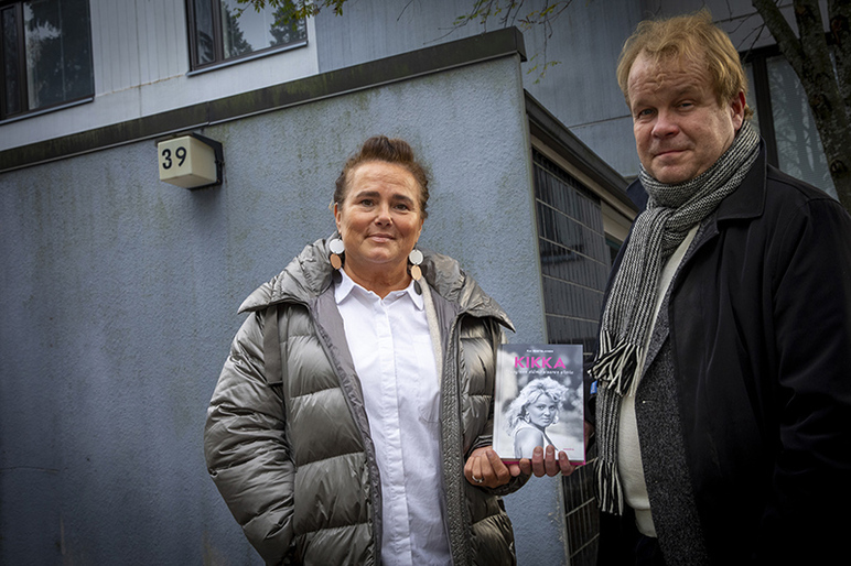 Puolivartalokuva kahdesta ihmisestä harmaan kerrostalon alakerran edessä. Toisella heistä on kädessään kirja.