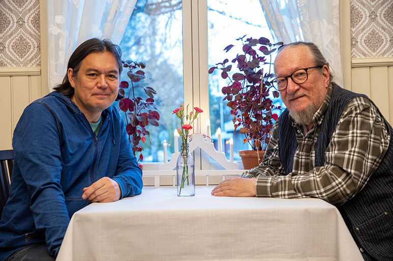 Kaksi miestä istuu ikkunan edessä olevan kotoisan oloisen pöydän ääressä. Pöydällä on kukkia lasipullossa, ja ikkunan edessä kynttelikkö ja kasveja.