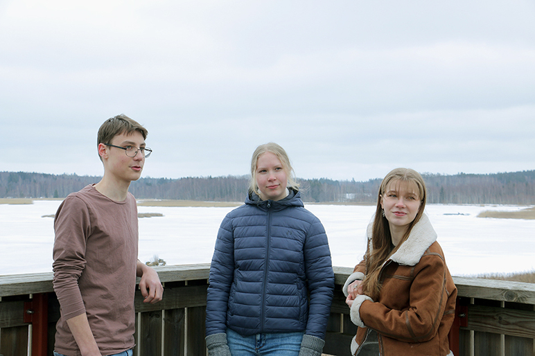 Kolme nuorta seisoo kevättalvisessa maisemassa kaiteen vieressä rannalla.