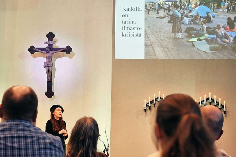 Nainen puhuu ristin alapuolella, ja edessä on ihmisiä selin. Seinällä näkyy iso dia, jossa on ihmisiä istumassa kadulla ja teksti Kaikilla on tarina ilmastokriisistä.