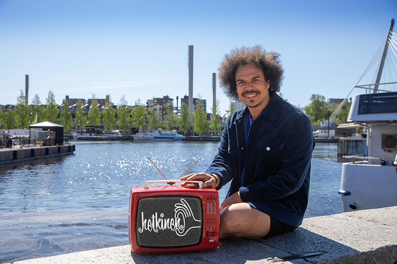 Naurava mies istuu shortsit jalassa veden äärellä. Vieressä reunavallilla on vanha minitelevisio, jossa lukee hetkinen.