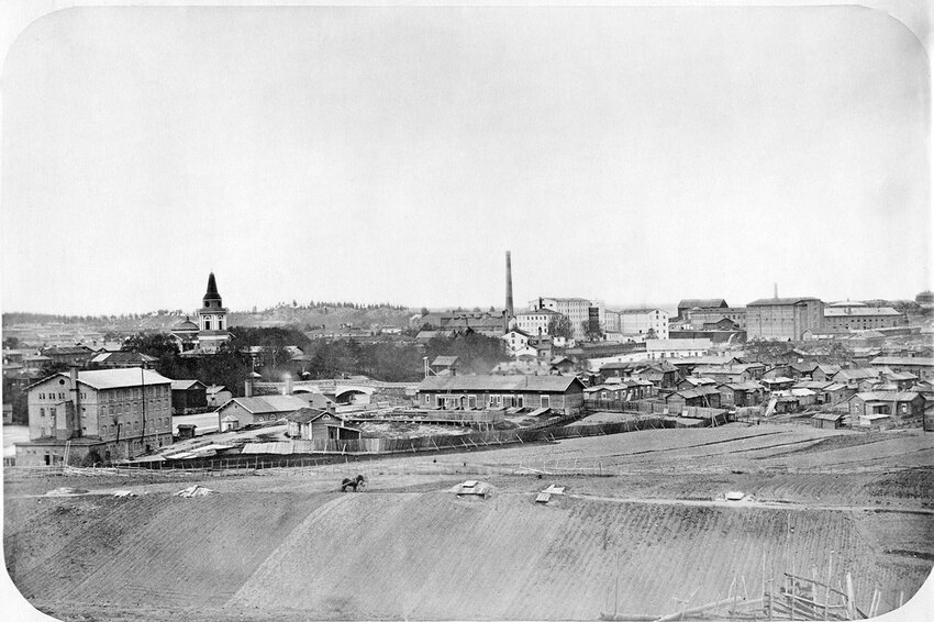 Yleisnäkymä Tampereelta vuodelta 1866, jossa Tammerkosken itäpuolella on puisia matalia majoja ja takana kohoaa Vanhan kirkon torni ja Finlaysonin tehtaan alueen rakennukset.