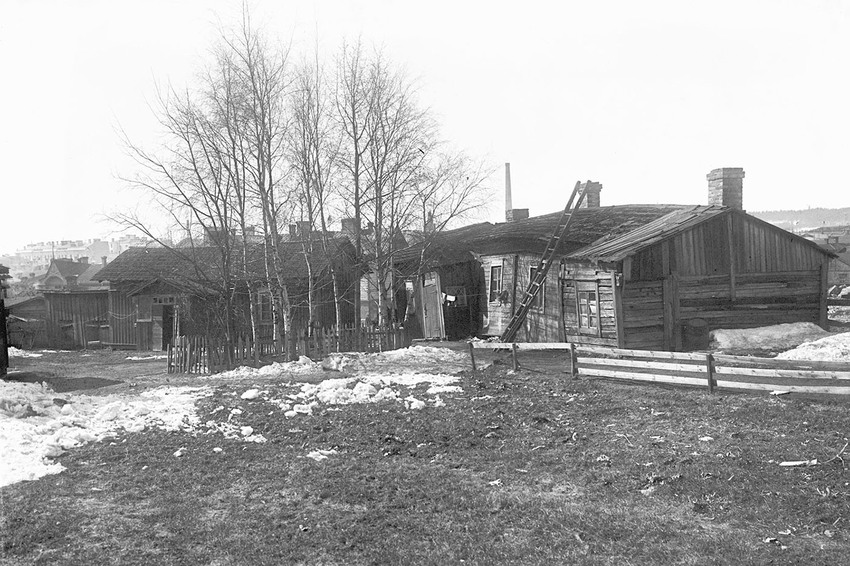 Mustavalkoinen kuva, jossa vanha 1900-luvun puutalo. Pihassa lehdettömiä puita ja sekä aitoja. Taloa vasten nojaa tikapuut.