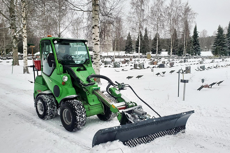 Pienkuormaaja työntää lunta lumisen hautausmaan käytävällä.