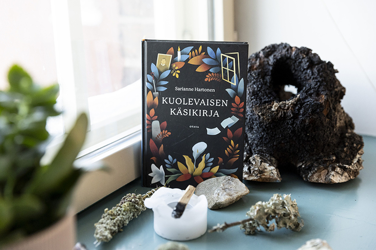 Ikkunan edessä on kirja, jäkäläisiä oksia ja kynttilä, jonka päällä on sytyttämiseen käytetty tikku.
