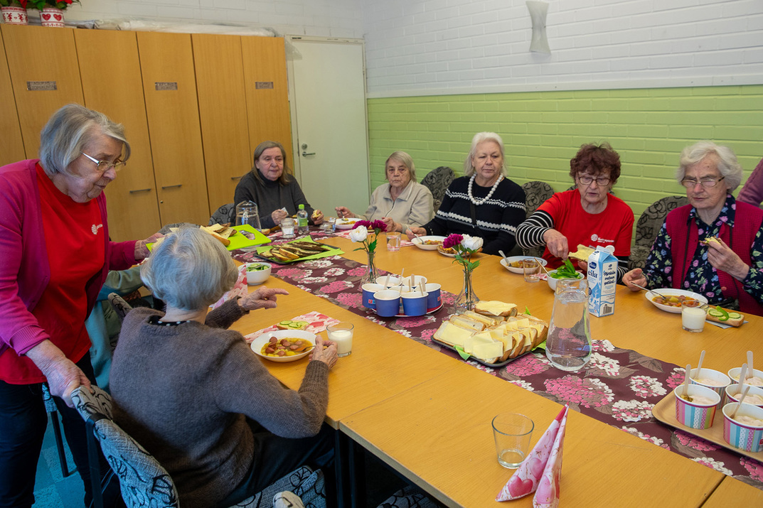 Ikäihmisiä istumassa pöydän äärellä ruokailemassa. Pöydällä kukkaliina sekä astioita ja ruokaa.