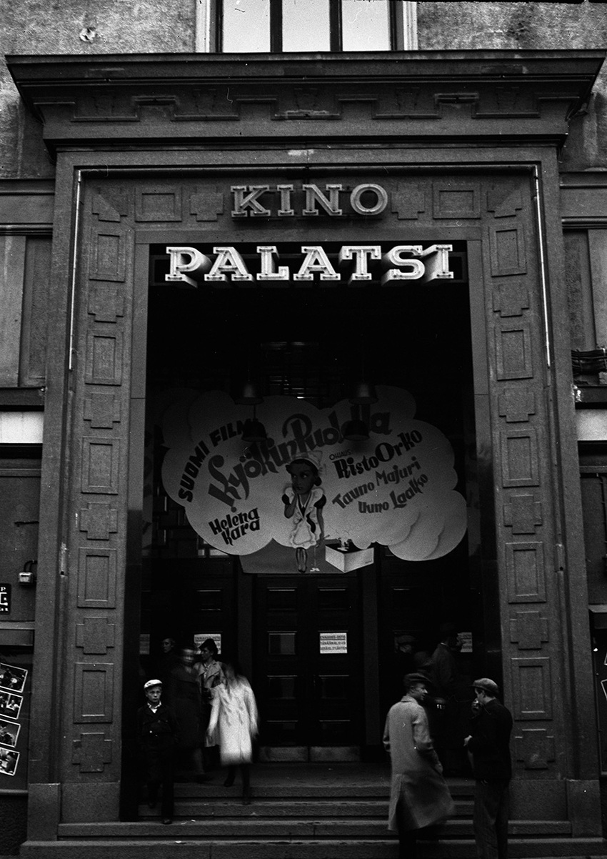 Mustavalkoinen kuva teatterin isosta ovesta ja rappusista. Ihmisiä kulkemassa rappusissa ja ovesta sisään.