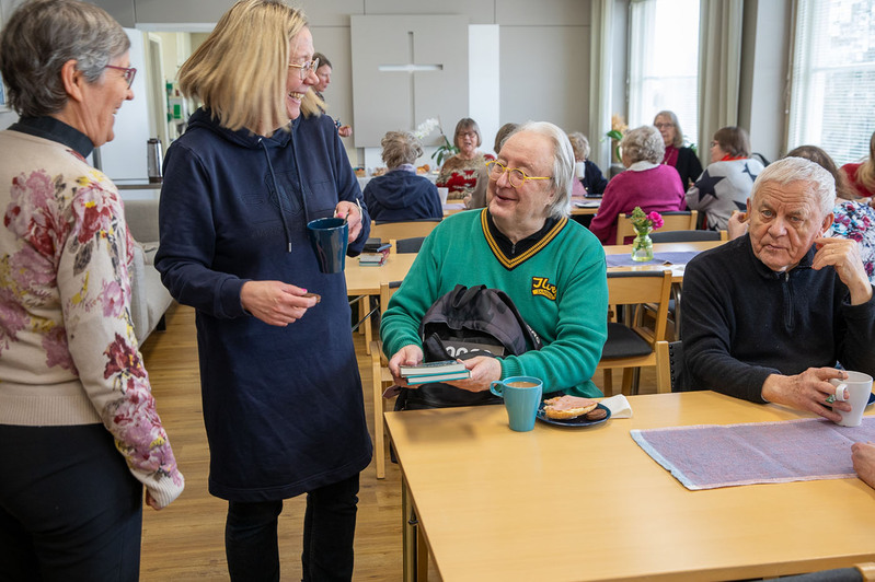 Mies istuu pöydän ääressä, edessään kahvikuppi ja syötävää. Mies pitää kädessään kahta kirjaa ja esittelee niitä edessään seisovalle naiselle. Molemmat hymyilevät. Taustalla eri ikäisiä ihmisiä seurakuntasalissa istumassa ja seisomassa pöytien ääressä.