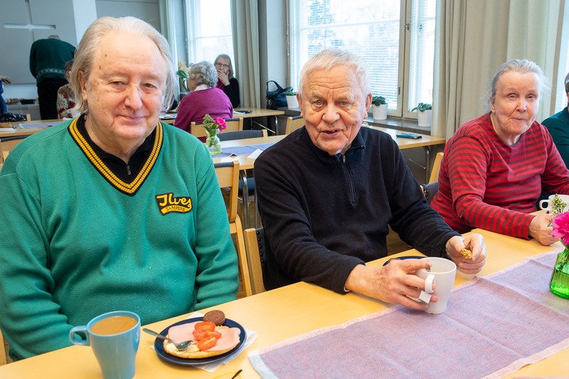 Kaksi miestä ja yksi nainen istuu pöydän ääressä kahvittelemassa.