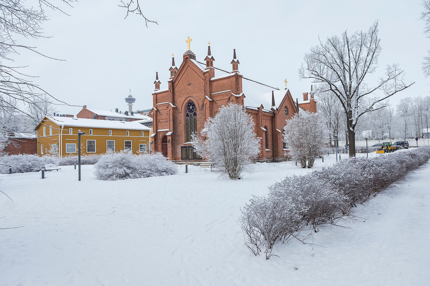 Punatiilinen kirkko talvimaisemassa. Taustalla keltainen puutalo. Puutalon katon yllä näkyy kerrostalojen kattoja sekä näkötorni.
