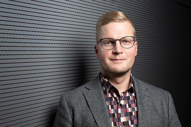 Ylävartalokuva henkilöstä, jolla on vaalea pukutakki ja ruudullinen paita.