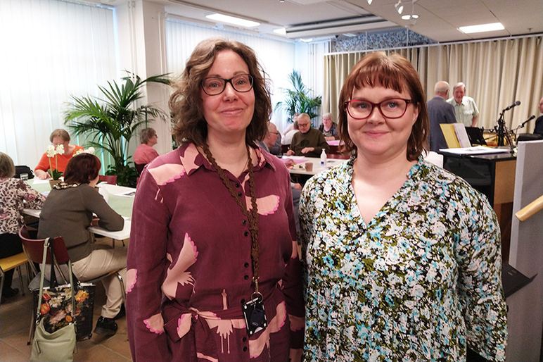 Ylävartalokuva kahdesta naisesta, joiden taustalla touhuaa paljon ihmisiä pöytien ääressä.