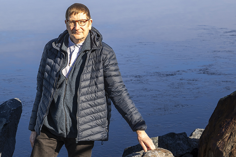 Kevyttoppatakkiin pukeutunut mies seisoo vesistön edessä ja pitää kättään rantakivellä.