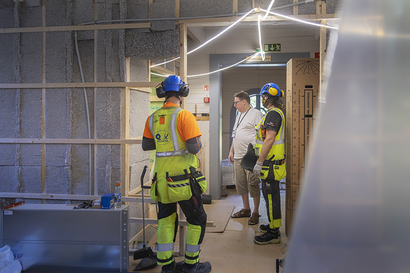 Kolme henkilöä, joista kahdella on päällään työhaalarit, katselee tilaa.