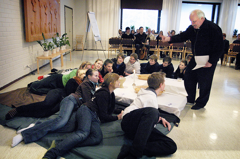 Vanhempi henkilö kävelee keskellä lattiaa heiluttaen kättään, ja toisessa kädessä on papereita. Lattialla makailee paljon nuoria.