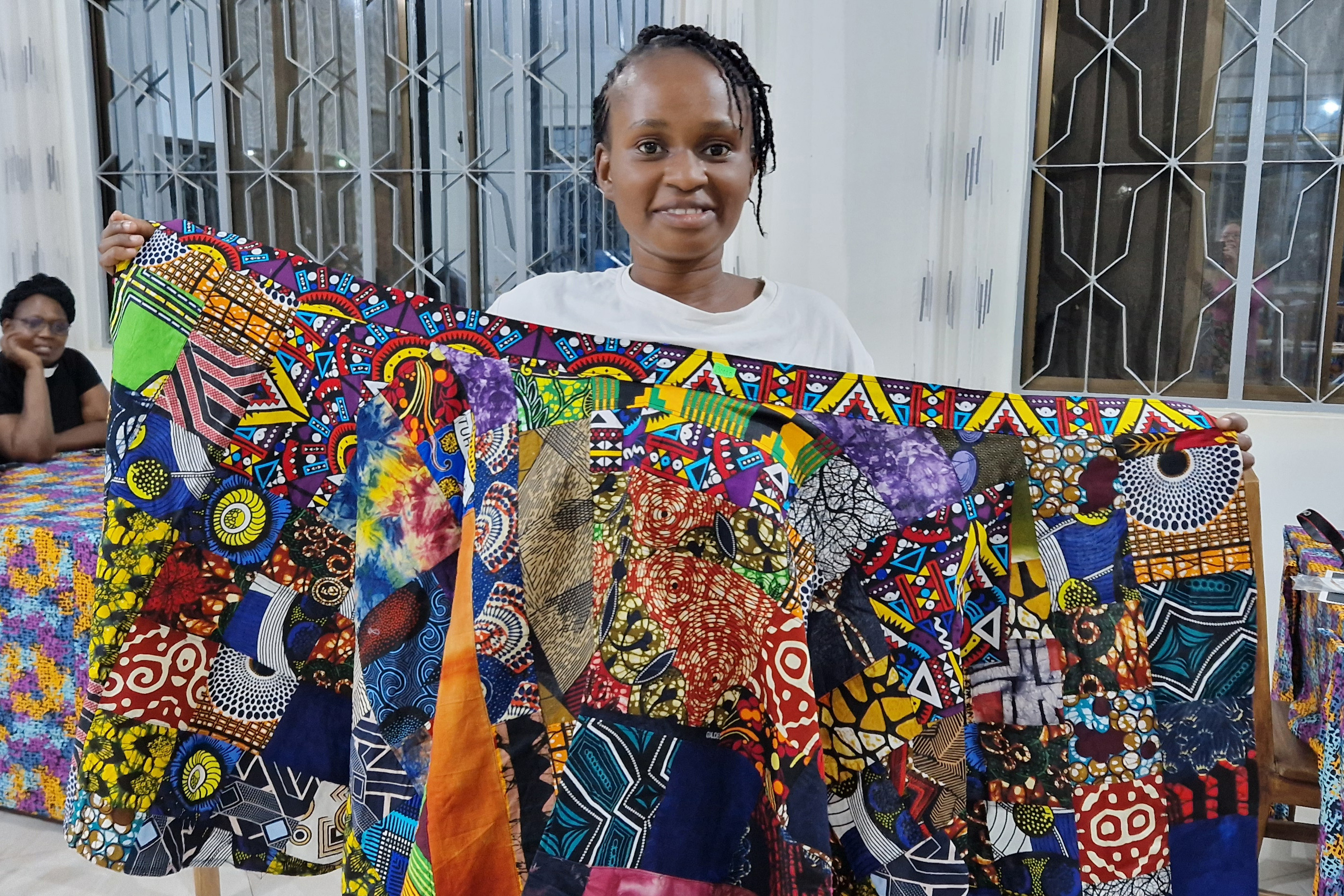 Nuori nainen esittelee isoa värikästä kangasta. Taustalla on nainen papin asussa.