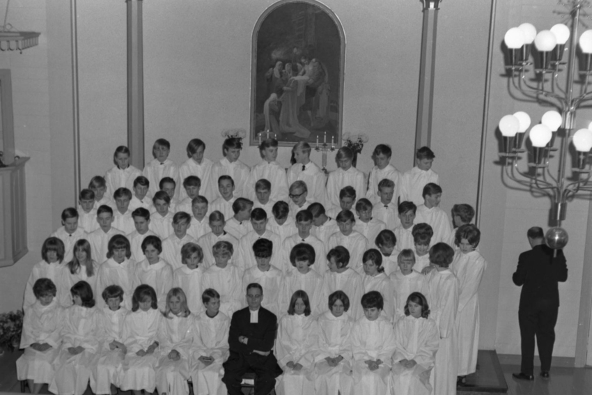 Mustavalkoisessa kuvassa joukko nuoria kirkon etuosassa ja heidän keskellään eturivissä pappi.
