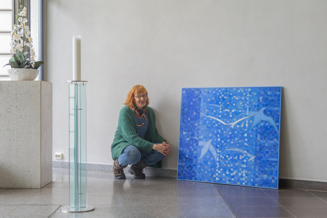 Nainen kyykistyneenä sinisävyisen taideteoksen viereen. Edustalla korkea, maasta asti oleva kynttilä ja sivustalla alttarin reuna, jolla kukkia.