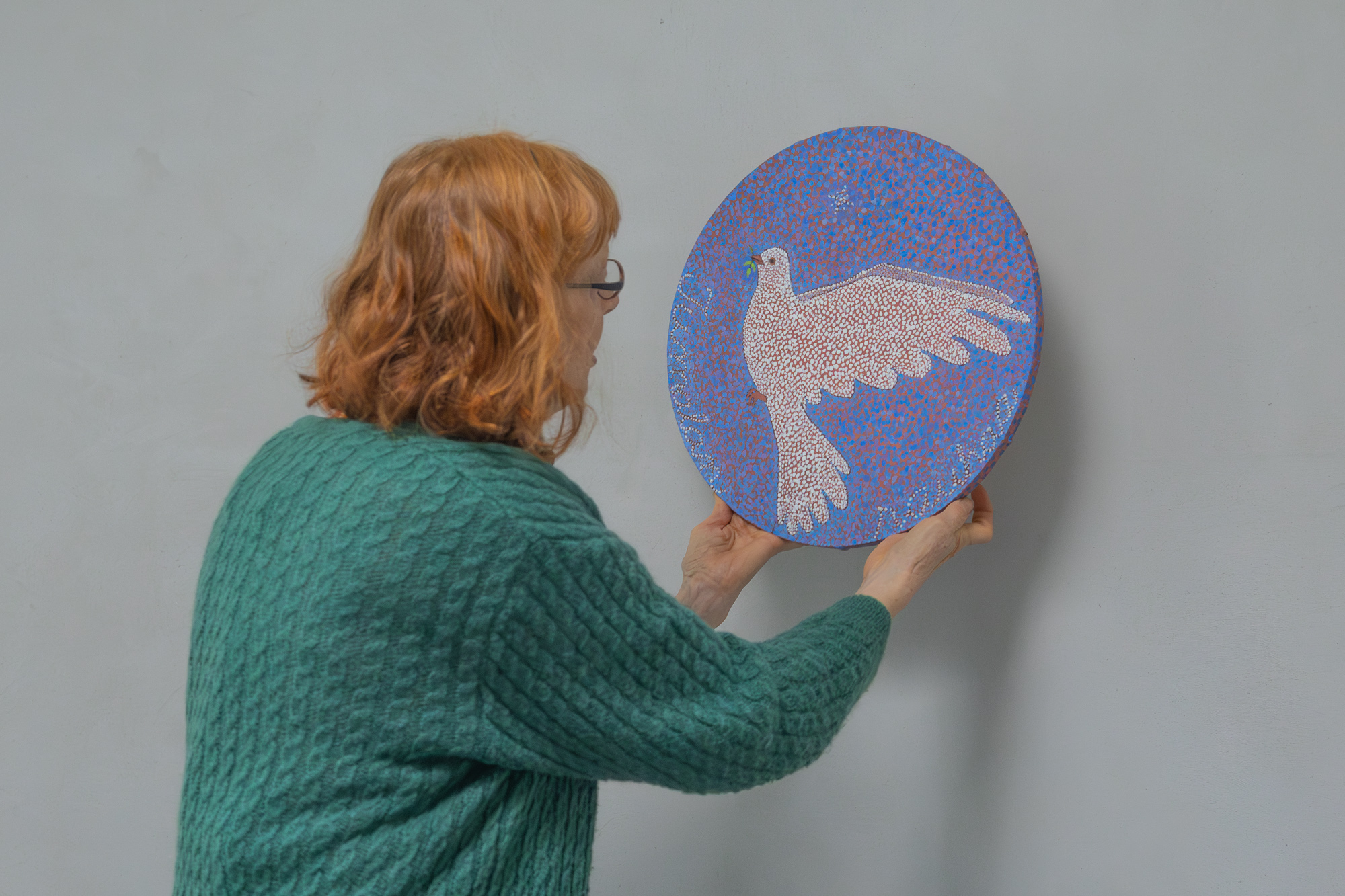 Nainen on selin ja asettelee seinäööe pyöreää taideteosta, joka on sinisävyinen ja siinä on kyyhkynen.