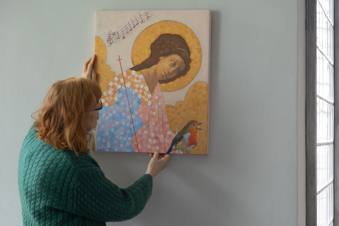 Nainen on selin kameraan ja asettelee seinälle taideteosta, jossa kuvattuna kullanhohtoinen enkeli sekä punarintainen lintu.