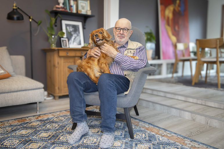 Mies istuu tuolissa ja rapsuttelee pitkäkarvaista lurppakorvaista koiraa, jolla on kieli ulkona suusta.