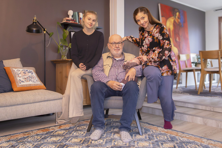 Keskellä huonetta tuolissa istuu mies pieni vauva sylissään. Hänen vierillään on kaksi naista, joilla molemmilla on toinen jalka tuolin käsinojalla.