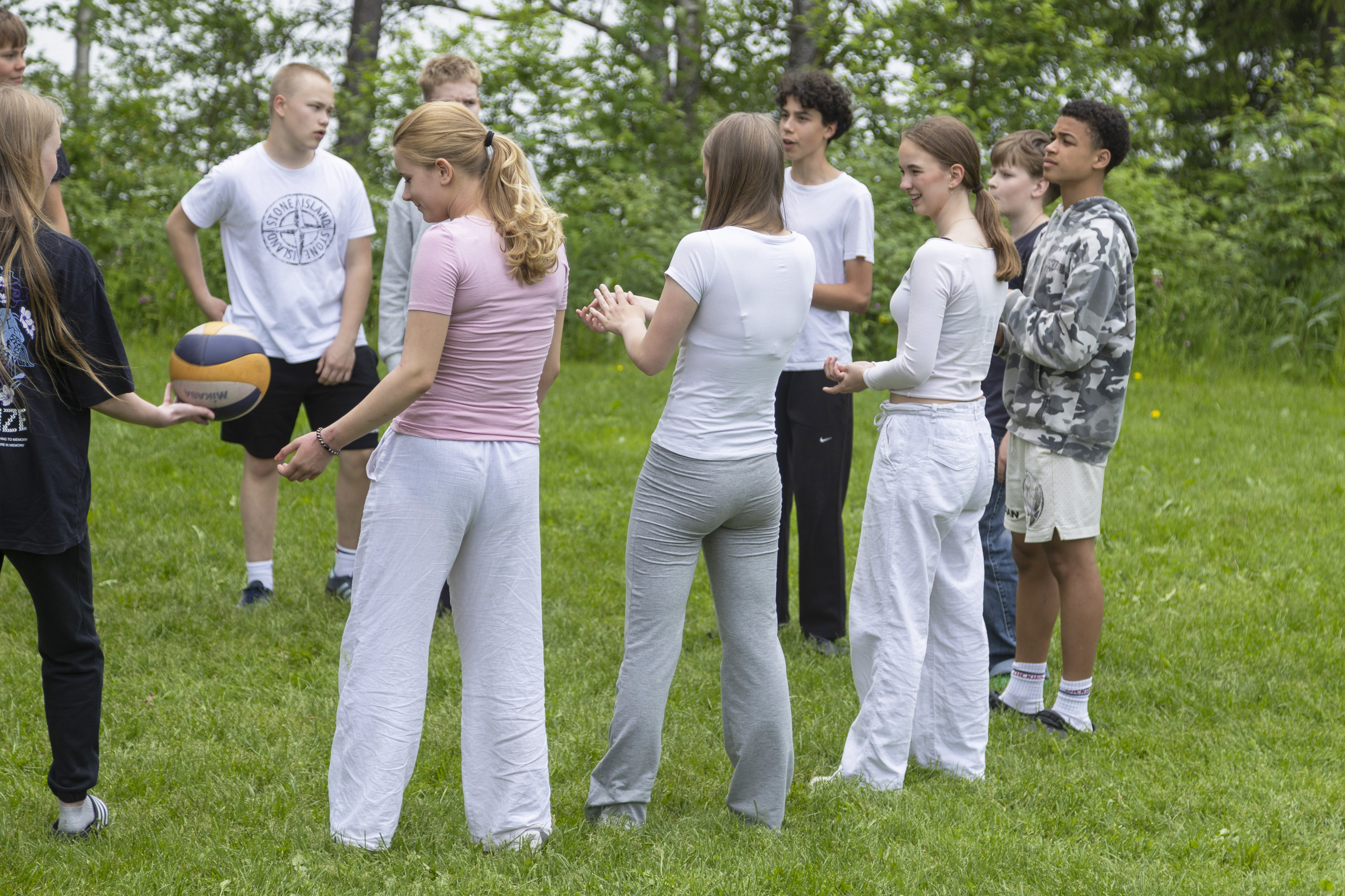 Joukko nuoria seisoo ringissä nurmikolla. Yksi heistä ojentaa toiselle palloa.