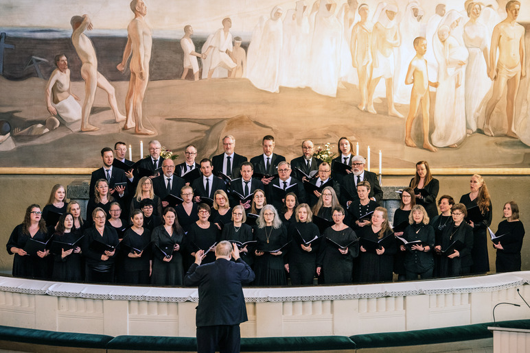 Kuoronjohtaja ison kuoron edessä kirkossa. Taustalla on iso maalaus, jossa alastomia tai kaapuun pukeutuneita hahmoja nousee maasta tai kävelee muodostelmassa.